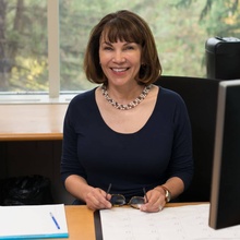 Headshot of Carin Crain smiling and seated at her desk in front of a bright window.