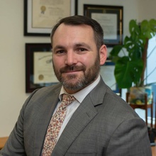 Headshot of Mihailis Diamantis from 2022 in a gray suit and flowered tie with several framed diplomas in the background.