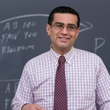 Faculty portrait of Andy Grewal, wearing a purple checkered shirt and lecturing a classroom in front of a blackboard.