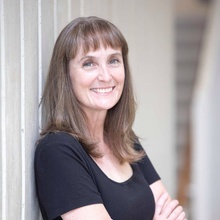 Professor Emily Hughes, wearing a black shirt and leaning against a white wood-paneled wall.