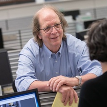 Faculty portrait of John Allen, wearing a light blue button-up shirt and having a conversation with someone out of frame.