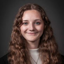 A woman with brown hair and blue eyes wearing a white shirt with a black sweater and a silver necklace smiling
