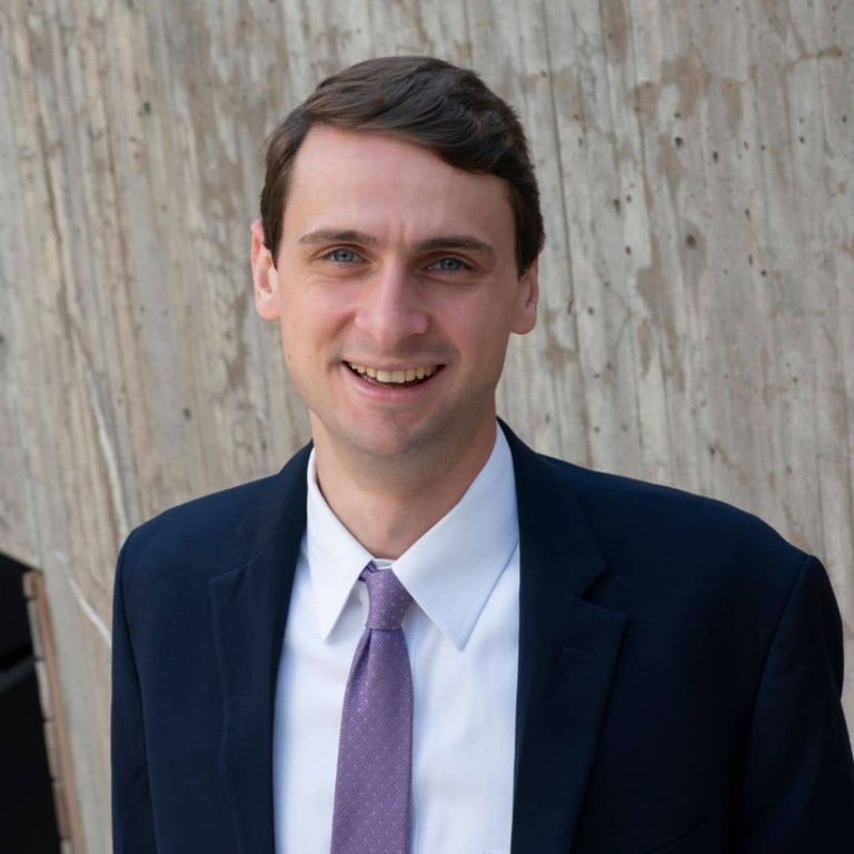 Faculty portrait of James Toomey, wearing a dark blue suit jacket, white shirt, and purple tie, smiling in front of the BLB portico wall.