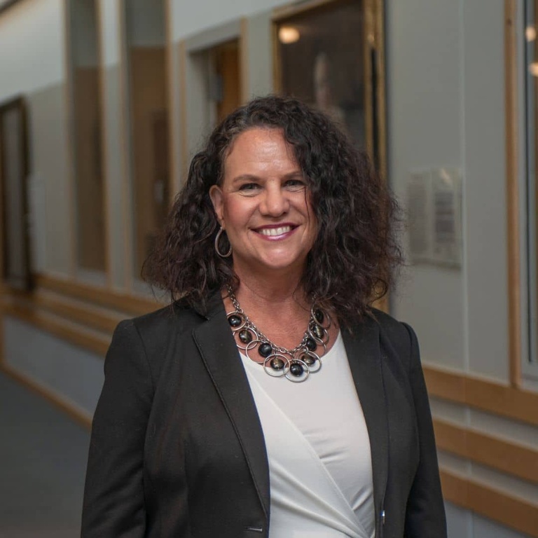 Headshot of Sara Clark, wearing a black coat and white shirt and smiling in the hallway of Boyd Law Building.
