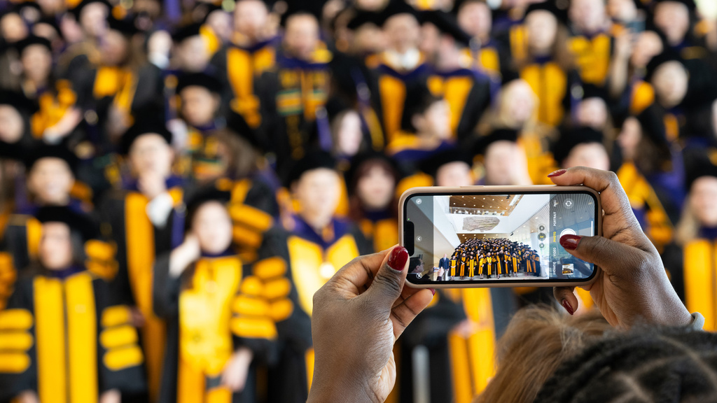 An audience member holding up a cell phone camera to take a picture of the graduating class, lined up on a staircase in Hancher Auditorium.